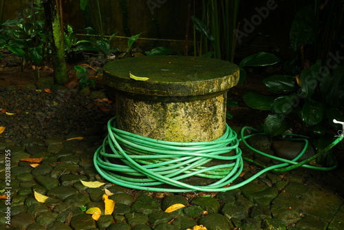 Weathered stone pedestal wrapped with a vibrant green rubber garden hose sits on a damp cobblestone path surrounded by lush tropical foliage in a quiet and humid backyard garden setting.