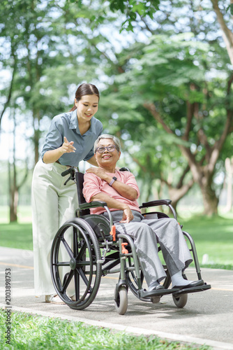 Young caregiver pushing older woman in wheelchair along path in park