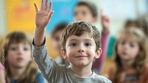 A young boy raises his right hand in a classroom setting, smiling and waving.