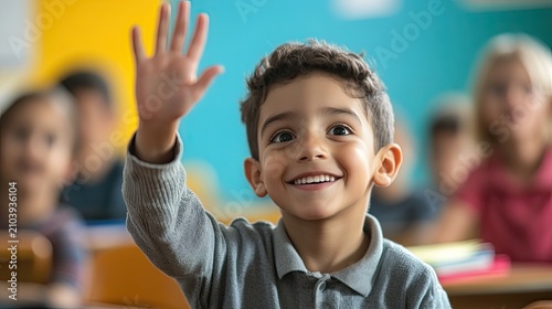 A young boy in a classroom raises his hand enthusiastically, smiling broadly.