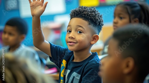 A young boy with curly hair, wearing a blue shirt with yellow stripes, raises his hand in a classroom setting.