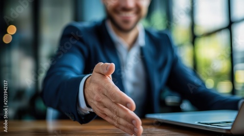 A businessman in a suit is extending his hand for a handshake, likely in a professional setting with a laptop and blurred office background.