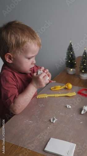 Toddler playing with air dry clay and making handmade holiday decorations at home. Three-year-old boy shaping air dry clay with toy tools at home. Cozy holiday atmosphere with miniature Christmas tree