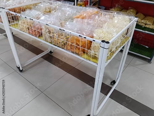 A retail display table filled with assorted packaged traditional crackers, chips, and dried snack foods in clear plastic bags inside a grocery or snack shop. The white wire basket display showcases a 