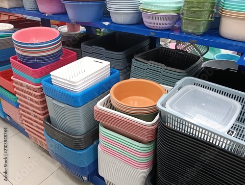 A retail store display featuring a wide variety of colorful plastic household storage containers, baskets, trays, tubs, basins, and colanders neatly stacked on shelves. The assortment includes storage