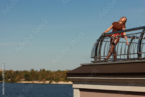 Full length portrait of a beautiful young woman with long light brown hair in a brown blouse and pants sitting on a metal railing over a city river