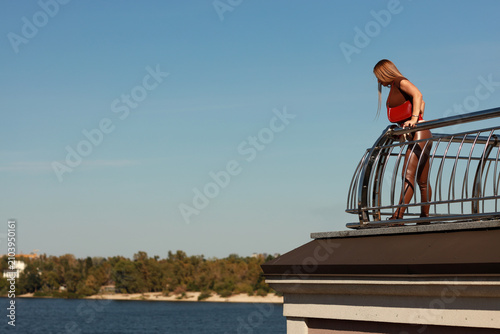 Full length portrait of a beautiful young woman with long light brown hair in a brown blouse and pants sitting on a metal railing over a city river