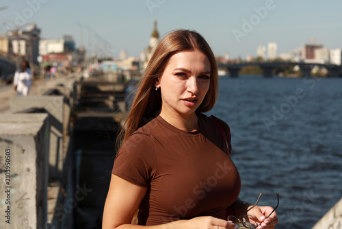 Close up portrait of a beautiful young woman with long light brown hair in a brown blouse against a blurred city river background in Kyiv