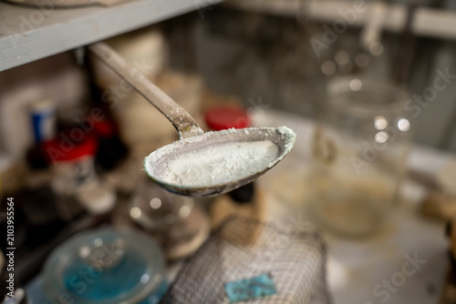 Konstfotografi Metal spoon filled with white enamel powder or flux for firing, held over a workshop table with refractory firebricks in the blurry background