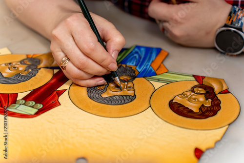 Close up of artist hands painting religious icon figures on a metal plate using hot vitreous enamel technique on a studio workbench.