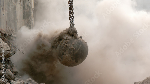 A wrecking ball swings into a building, breaking down the structure. Dust and debris fill the air at the demolition site during daytime hours