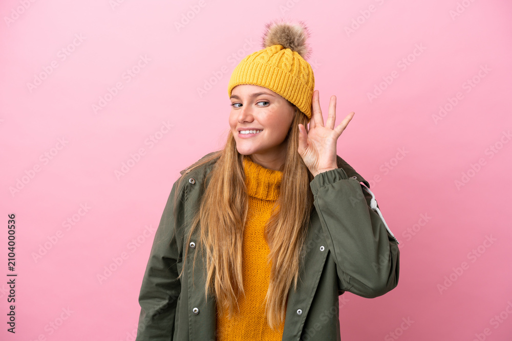 Obraz premium Young blonde woman wearing winter jacket isolated on pink background listening to something by putting hand on the ear