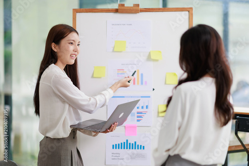 Businesswoman standing presenting project data and sales growth charts on a whiteboard during a corporate office meeting, discussing strategy with a colleague