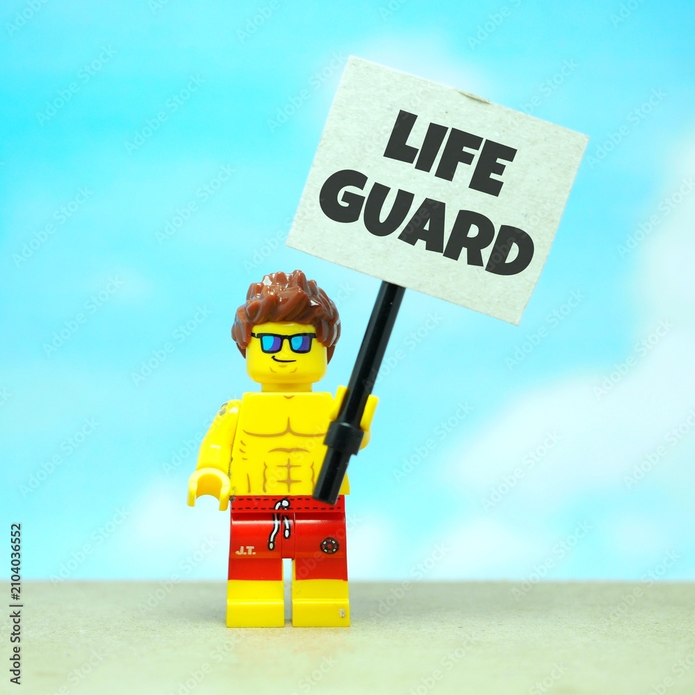 Fototapeta premium Indonesia - July 12, 2026 : Mini toy of action figure at outdoor with blur background. Miniature photography concept design. Minifigure of man male people hold LIFE GUARD text at poster banner.