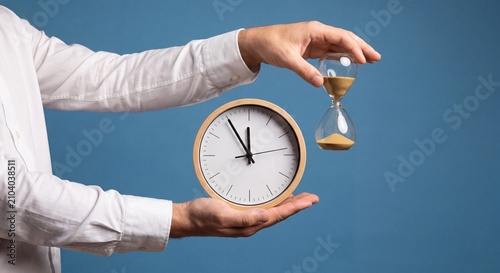 Business professional holding wooden wall clock and glass hourglass against solid blue background studio shot with shirt holding circular captures person white