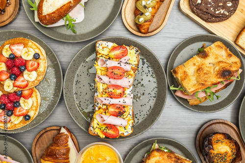 Top-down view of a breakfast spread featuring fruit pancakes, scrambled egg and ham toast, sandwiches, pastries, and orange juice on gray plates and wooden boards.