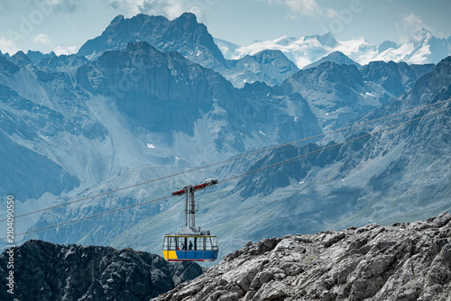 A colorful cable car travels above rugged alpine terrain, surrounded by dramatic rocky peaks in the Swiss alps, snow-covered mountains, and glaciers in a breathtaking high-altitude landscape.