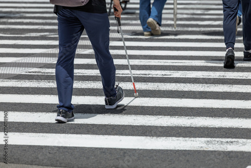 A visually impaired person using a white cane while walking across a marked city crosswalk in daylight as other blurred pedestrians walk nearby.