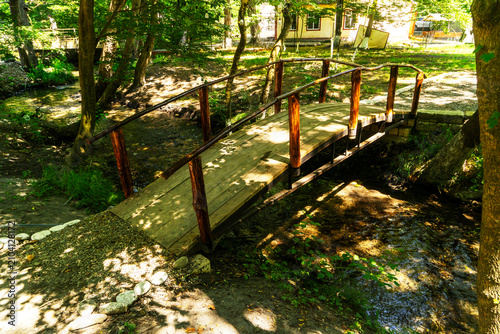 A picturesque wooden bridge over a clear forest stream in a green park. A cozy nature trail surrounded by tall trees and sunlight creates an atmosphere of tranquility and harmony. Photography is ideal