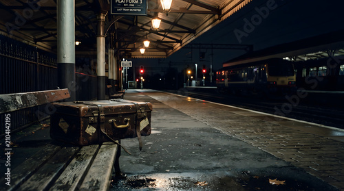 Vintage leather suitcase left on a wet bench at an empty train station platform at night under warm glowing lights