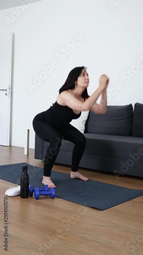 Young woman performs controlled deep squats on an exercise mat in a bright living room, promoting lower body strength, balance, mobility, wellness, and an active lifestyle.
