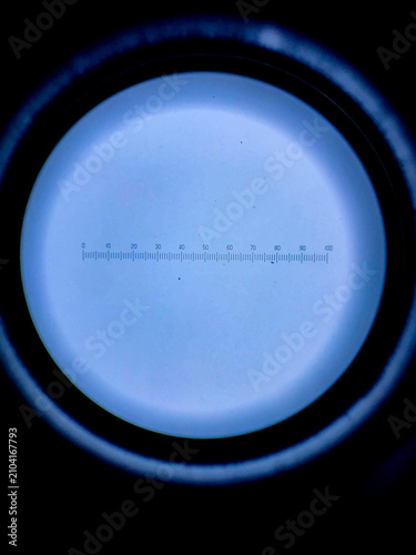 Circular microscope field of view showing an engraved measurement scale and crosshair reticle used for microscopy calibration and optical metrology.