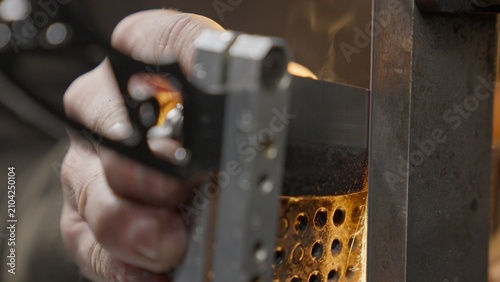Fotografie A blacksmith sharpens a wet knife blade on a belt grinder, producing metal sparks, steam, and smoke