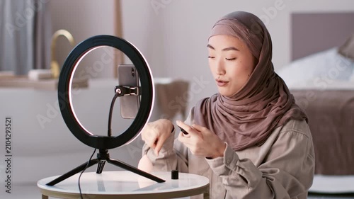 Focused Young Asian Woman Filming Makeup Tutorial With Smartphone In Front Of Well-lit Ring Light, Sitting At Table In Modern Home Setting. Creative Content Creation Concept.