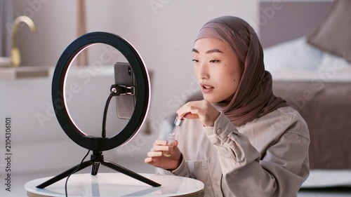 Focused Young Asian Woman Applying Makeup With Brush In Front Of Ring Light At Home, Sitting At Table. Indoor Setting With Natural Light, Engaged In Beauty Routine, Self-care Concept.