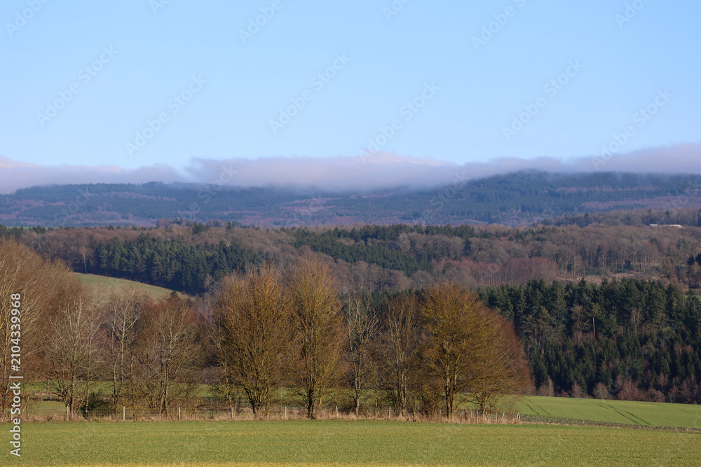 Fototapeta premium Wide landscape view of the forested Soonwald ridge with farmland and low clouds near Schönborn in Rhineland-Palatinate, Germany.