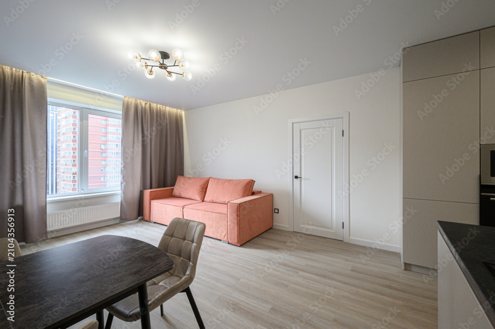 Fototapeta premium compact studio apartment features a warm orange sofa, a dining set, a wall‑mounted TV, neutral curtains, and light wood flooring, offering a bright, minimalist living space