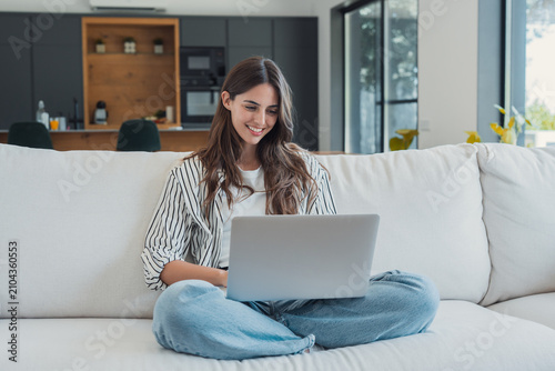Фотография Smiling young woman sits cross-legged on a white sofa using a laptop in a bright modern living room, portraying remote work, online study, leisure browsing and a comfortable home office lifestyle