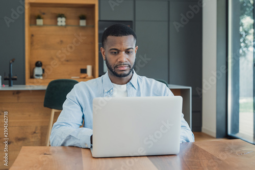 Slika na platnu A young man concentrates on his laptop at a modern kitchen table, working remotely and managing tasks in a bright, minimalist home office environment with natural light and a calm mood