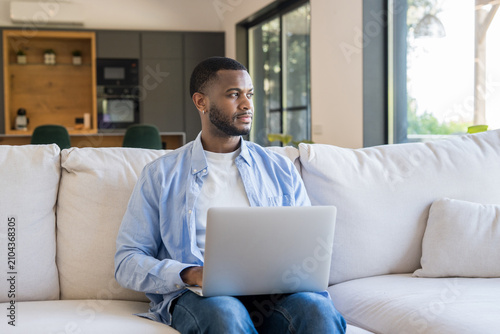 Foto Young man sits on a bright living room couch with a laptop, working remotely and gazing out the window, conveying focus, calm and a modern home office lifestyle