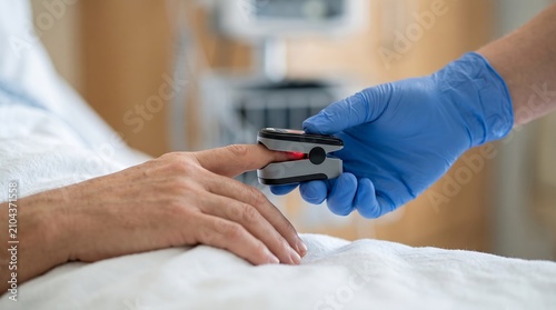 A healthcare professional in blue gloves places a pulse oximeter on a patient's finger in a hospital bed.