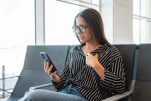 Foto Woman in glasses sitting in an airport waiting area anxiously checking her phone