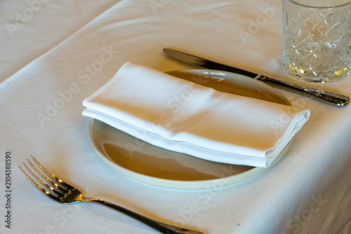 A neatly folded cloth napkin rests on a ceramic plate beside a knife, fork and clear drinking glass.