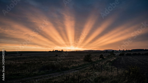 Radiant golden sun rays shining through dramatic clouds over a vast rural landscape during sunset