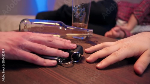 Empty glass bottle from alochol beverage beside car key fob and resting hands on wooden tabletop indoor living room. A male sober hand takes away the car keys to prevent a drunk person from driving