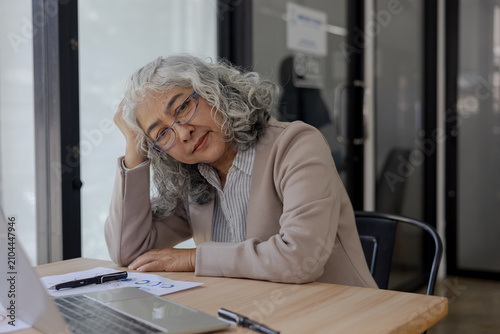 Foto Tired Senior Businesswoman Resting Head on Hand at Office Desk with Laptop and D