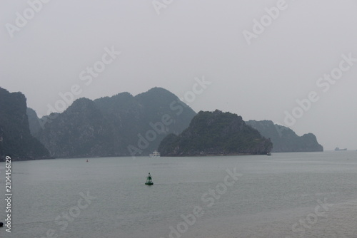 Green buoy floats in calm water near misty islands and distant ship.