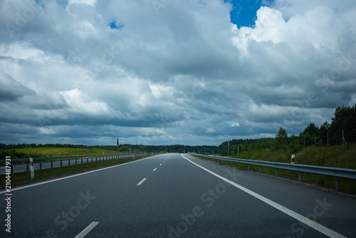 First-person perspective from a car driving on an empty asphalt highway. A modern expressway with a bridge overpass under a dramatic, cloudy stormy sky. No other traffic.