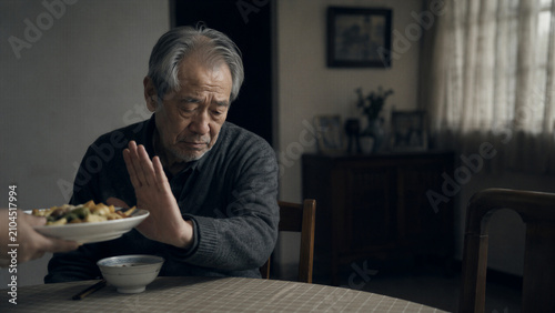 Elderly man refusing offered meal at dining table in kitchen