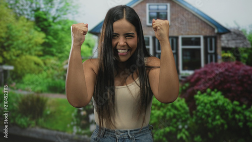 Fotografie Woman raising fists and smiling with bare arms visible in front of a residential building, celebrating a win and pumping her fists; joy victory