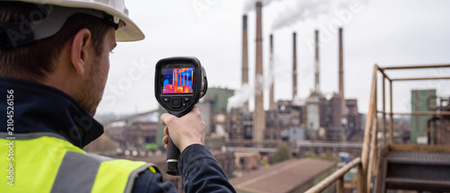 Industrial worker using a thermal imaging camera to inspect factory emissions and heat loss from industrial smokestacks