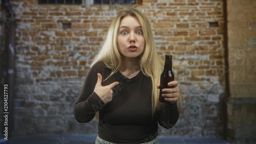 Foto Woman points finger to chest while holding a beer bottle in front of a brick building with a startled expression; confusion