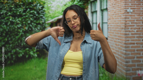 Photography Woman wearing glasses and yellow tank top showing thumbs up and thumbs down with hands in front of building brick wall and lawn; ambivalence