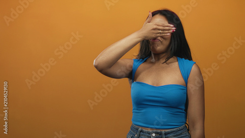 Canvas Print Woman covering eyes with hand, palm over face, wearing blue strap top and jeans, long brunette hair visible against orange studio wall; shyness