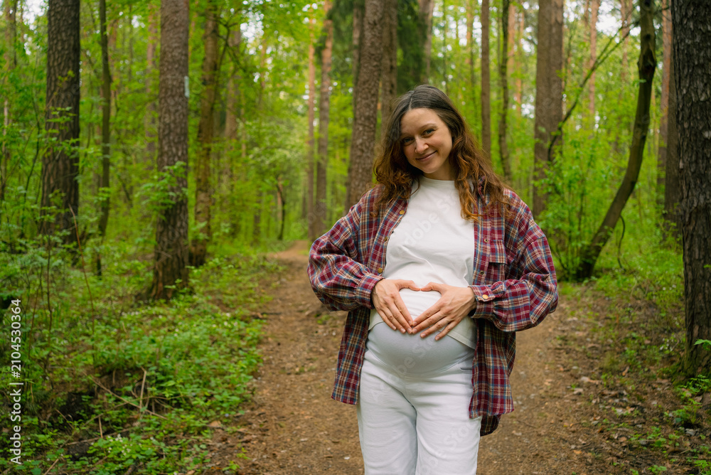Fototapeta premium Pregnant woman standing on a forest path, forming a heart shape with her hands on her baby bump, smiling and feeling happy, symbolizing new beginnings and awaited arrival