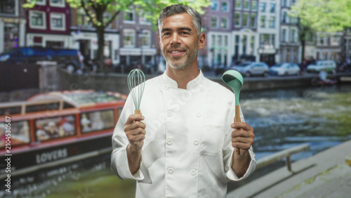 Φωτογραφία Man chef in white coat smiles and holds a whisk and ladle with bare hands beside a city canal street waterfront; confidence